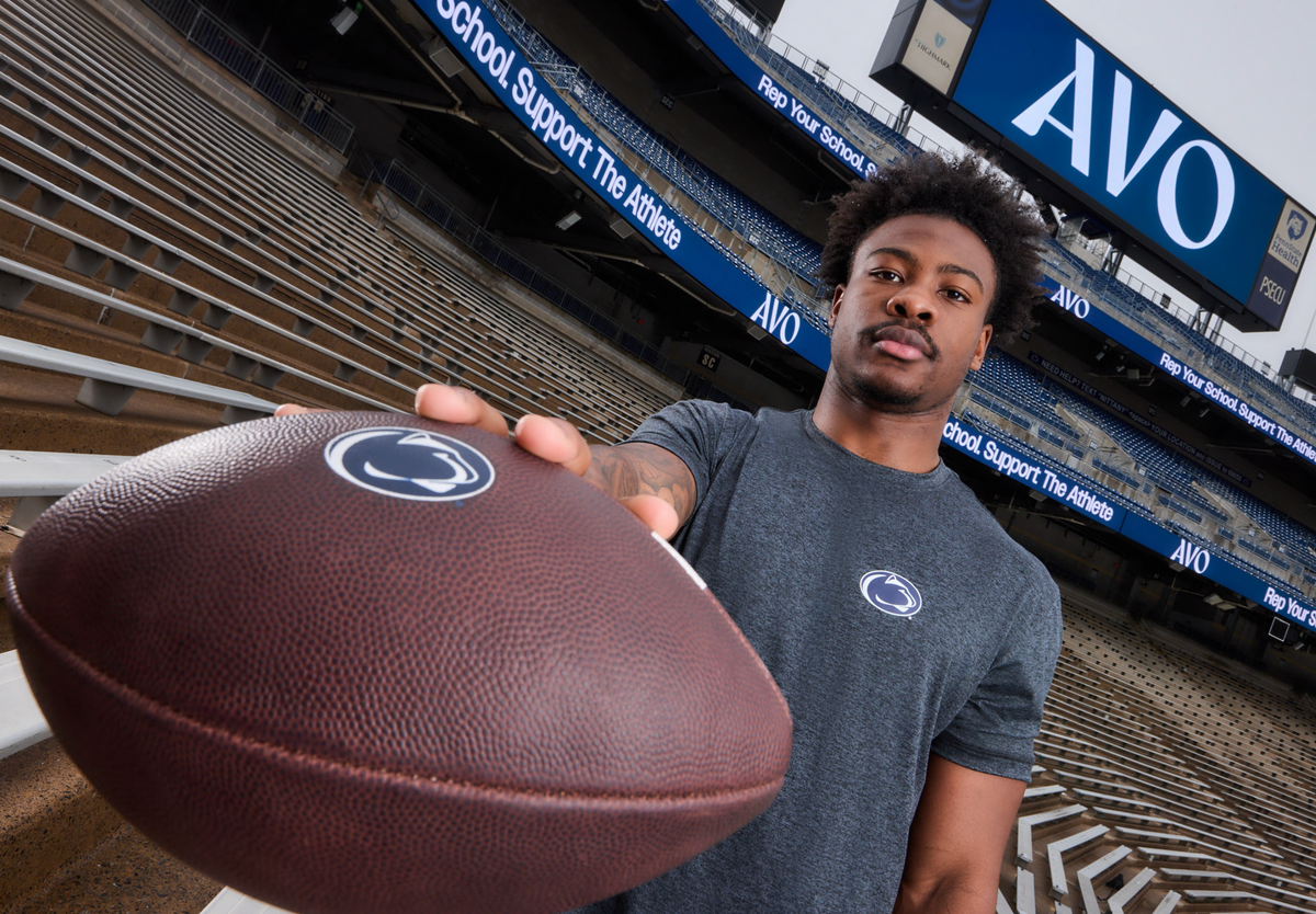 A man in a gray Penn State Nittany Lions t-shirt holds a brown football with the Penn State logo, posed in front of an empty stadium with AVO signage in the background.