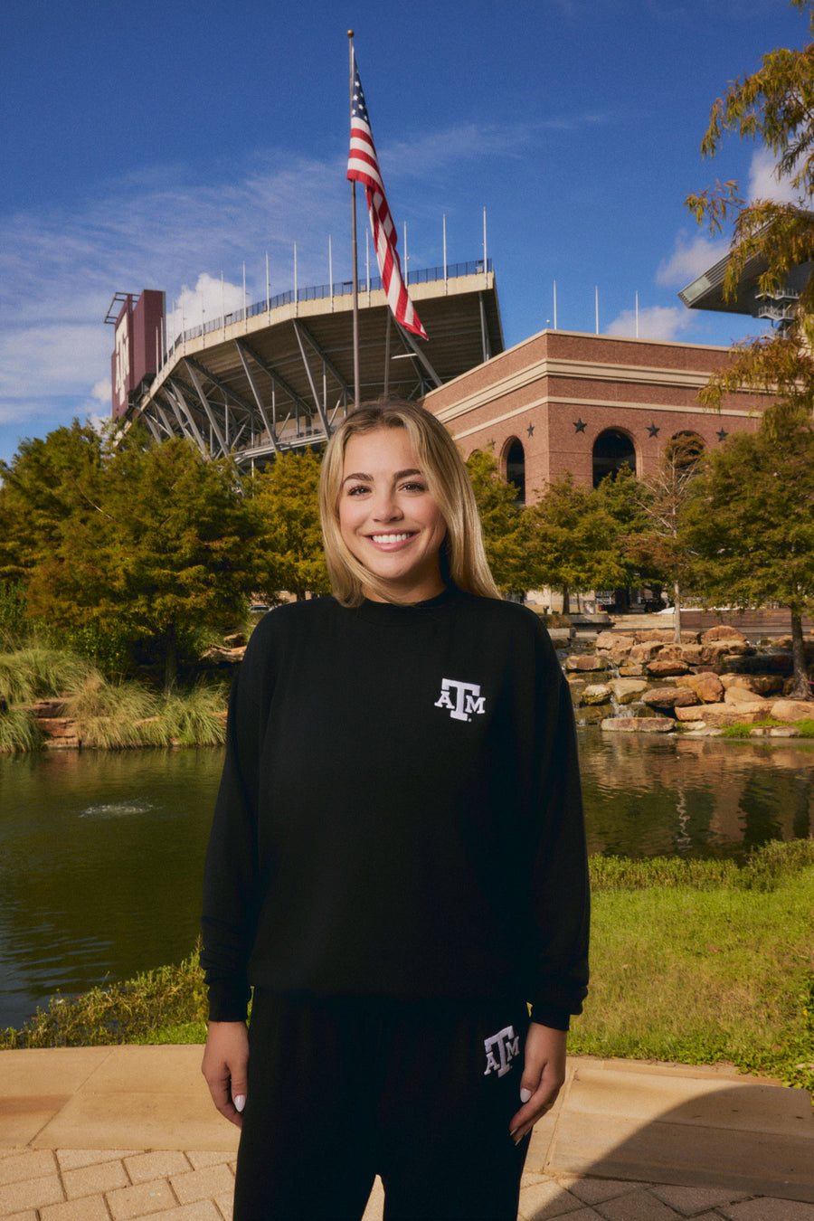 A smiling woman stands outdoors in front of a stadium and pond, wearing a black long-sleeve sweatshirt and matching black sweatpants with a white Texas A&M logo on the chest and leg.