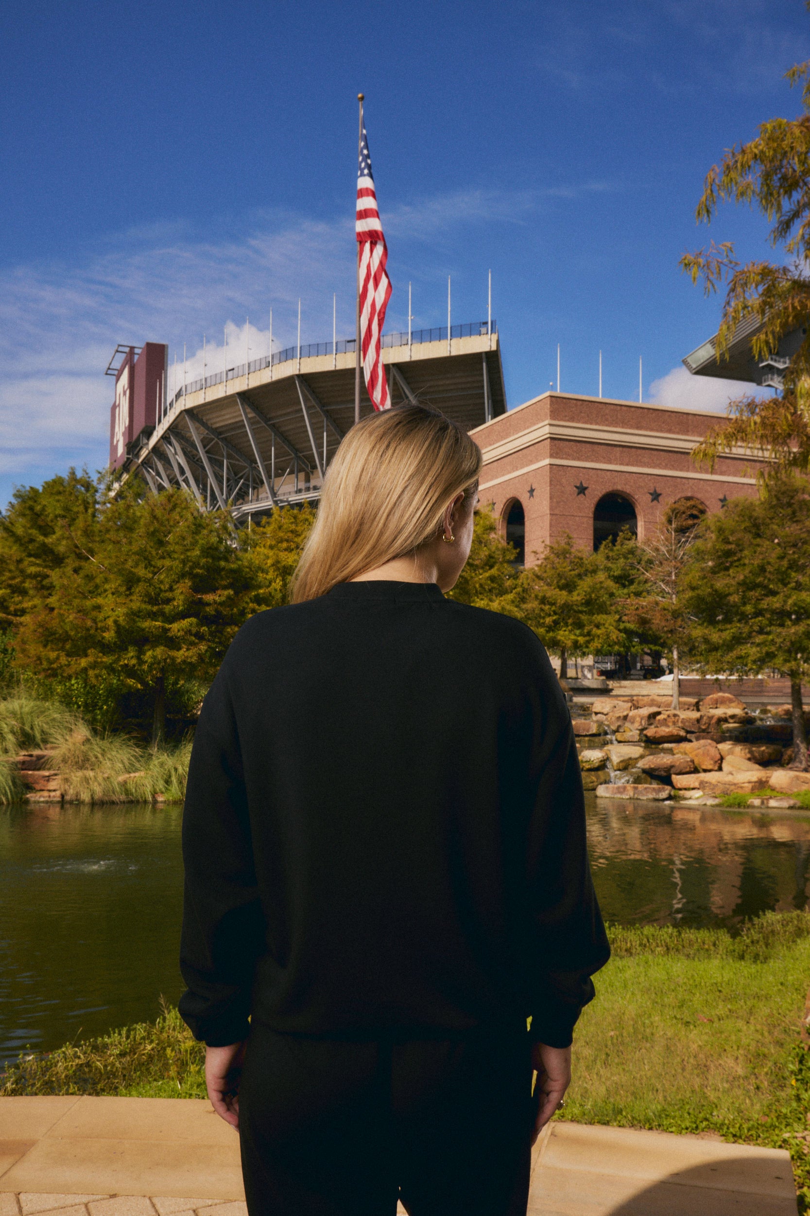 A woman with blonde hair is seen from behind, wearing a black sweatshirt and black pants while standing outdoors in front of a stadium and an American flag under a blue sky.