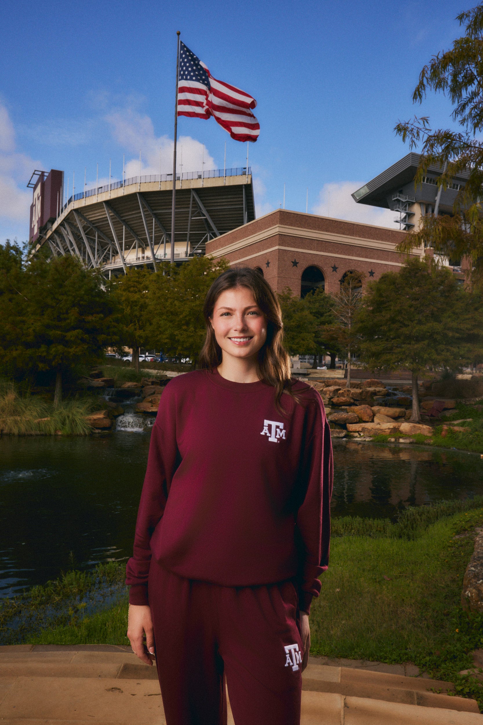 A woman stands outdoors in front of a stadium and pond, wearing a matching maroon sweatshirt and sweatpants set with white 'ATK' lettering. An American flag is visible in the background.