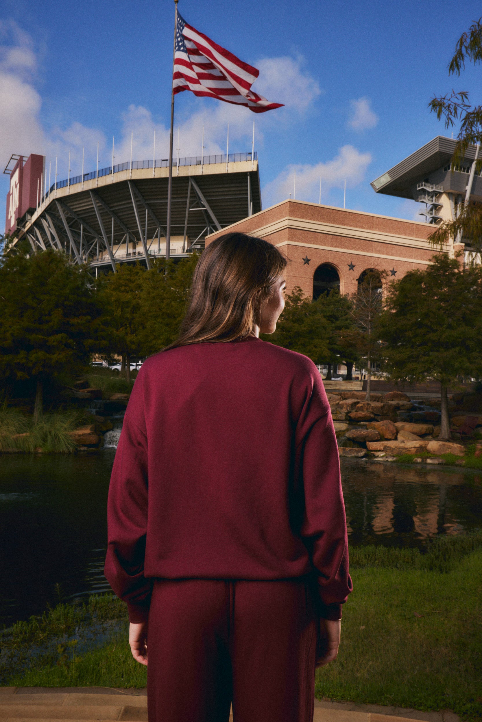 A woman viewed from behind wearing a matching burgundy sweatshirt and sweatpants set, standing outdoors in front of a stadium and pond with an American flag in the background.