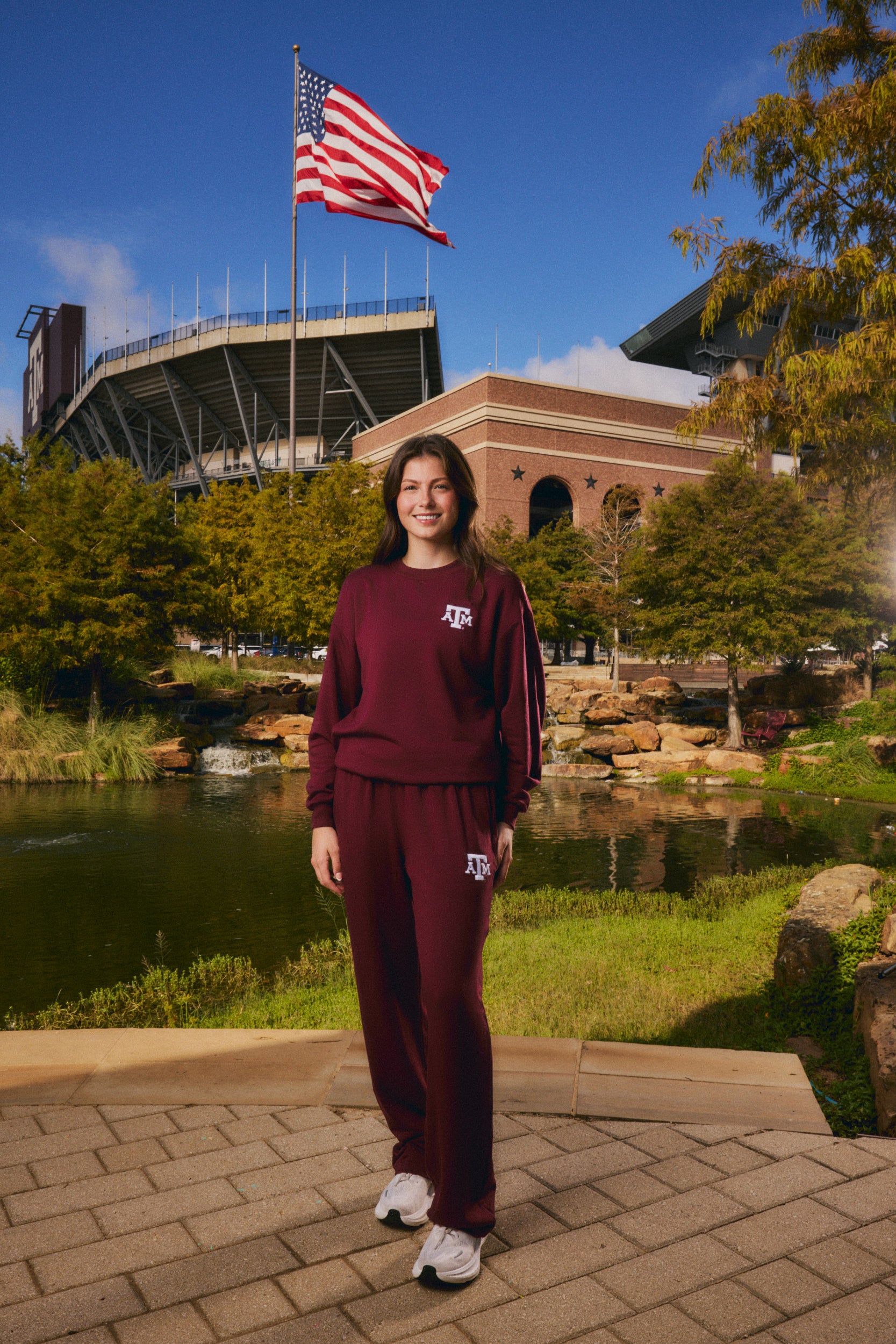 A woman stands outdoors in front of a stadium and pond, wearing a matching burgundy sweatshirt and sweatpants set with a white Texas A&M logo. An American flag flies above, with trees and blue sky in the background.
