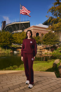 A woman stands outdoors in front of a stadium and pond, wearing a matching burgundy sweatshirt and sweatpants set with a white Texas A&M logo. An American flag flies above, with trees and blue sky in the background.