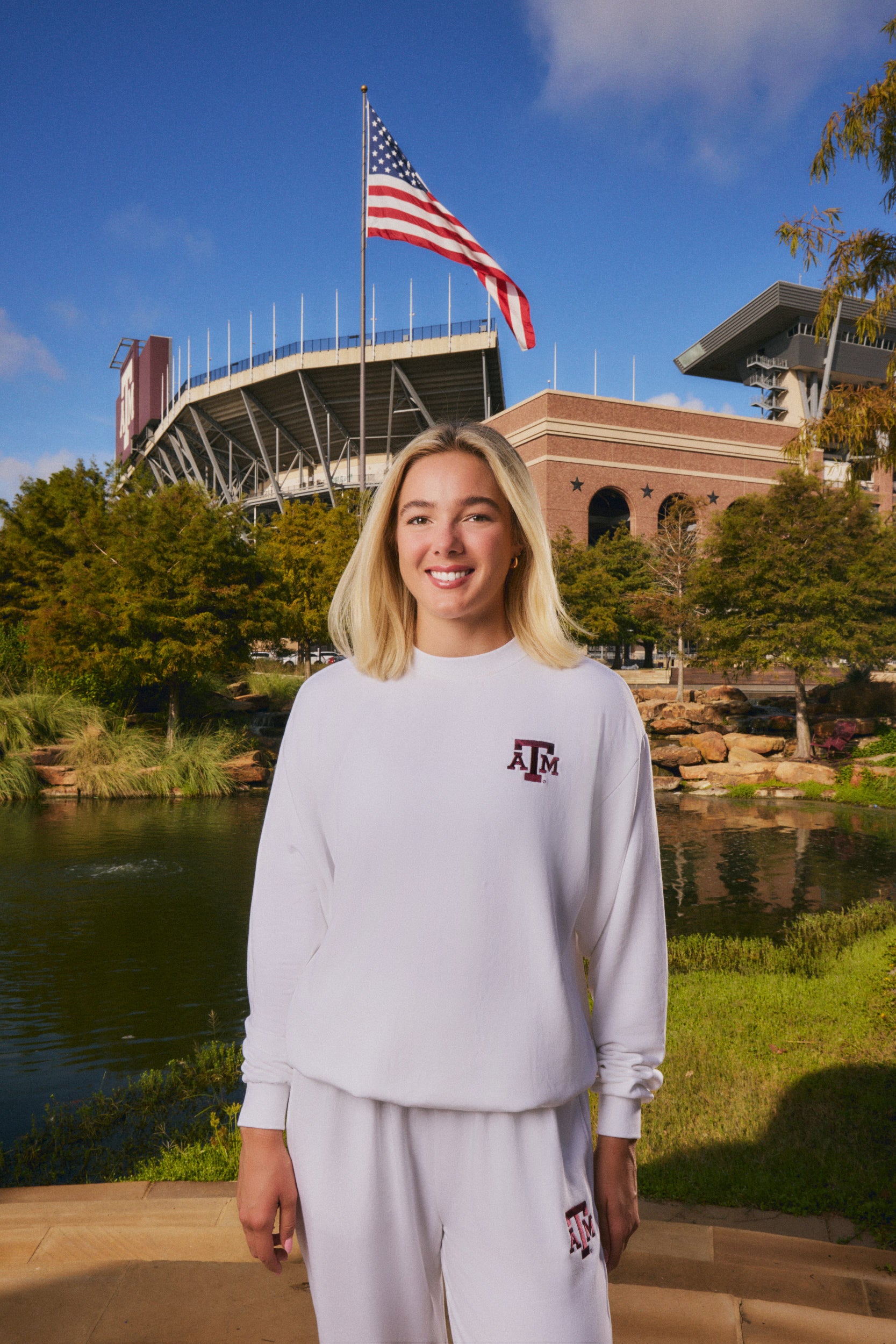 A smiling blonde woman stands outdoors in front of a stadium and pond, wearing a white long-sleeve sweatshirt and matching sweatpants with Texas A&M collegiate branding.