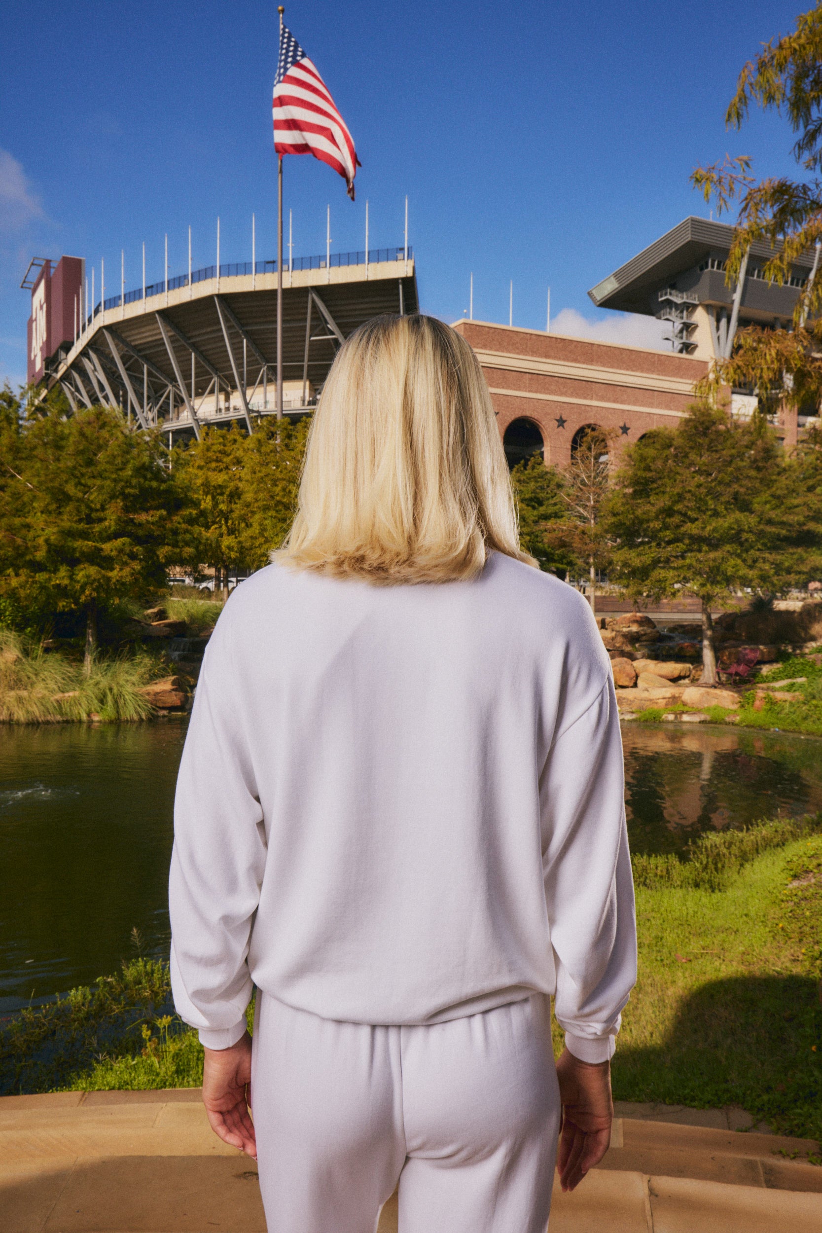 A woman with blonde hair is seen from behind, wearing a matching white sweatshirt and sweatpants set, standing in front of a stadium and pond with an American flag in the background.