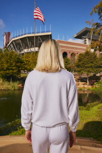 A woman with blonde hair is seen from behind, wearing a matching white sweatshirt and sweatpants set, standing in front of a stadium and pond with an American flag in the background.