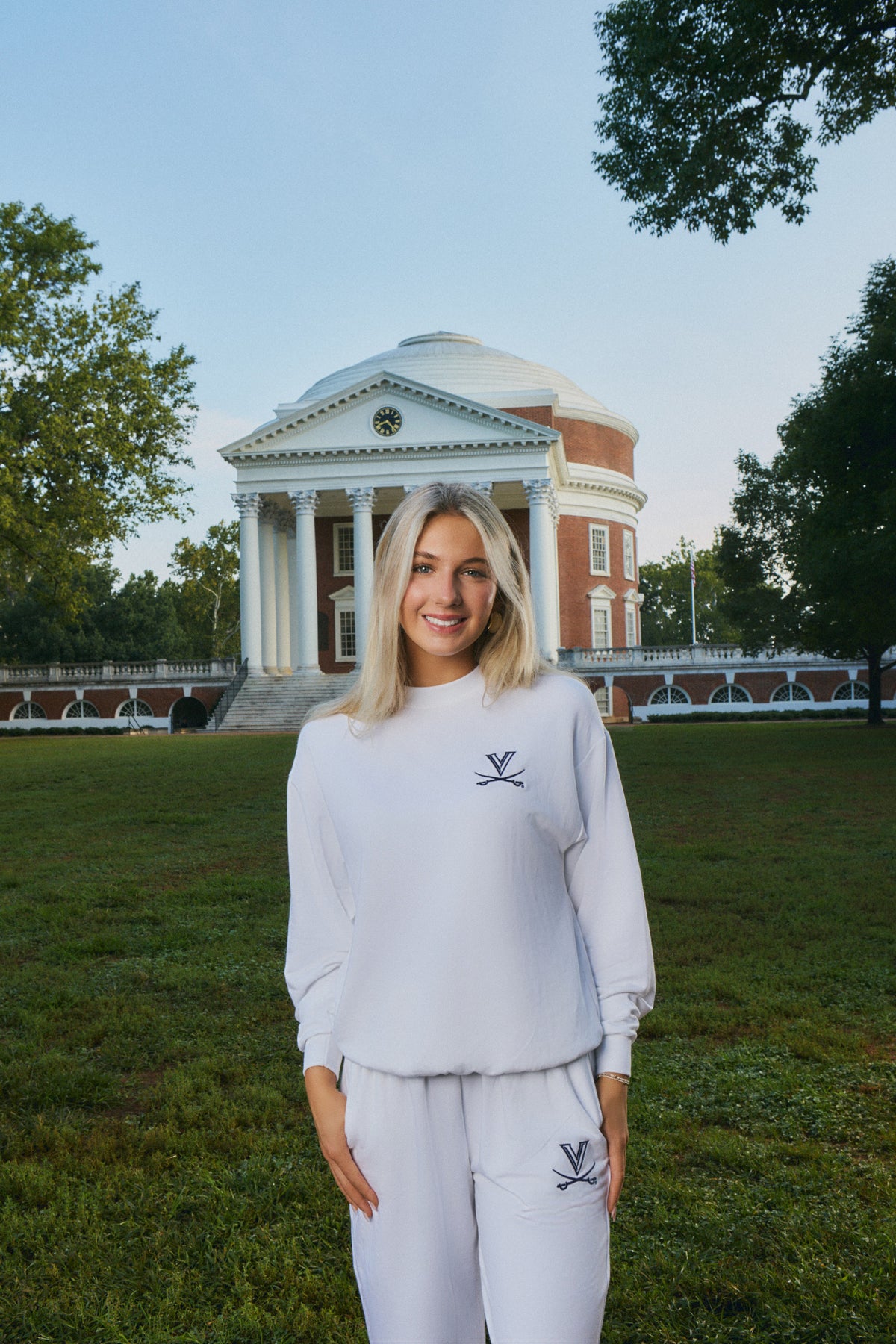 A woman stands outdoors on a grassy lawn in front of a large classical building, wearing a matching white sweatshirt and sweatpants set with a small dark embroidered logo on the chest and thigh.