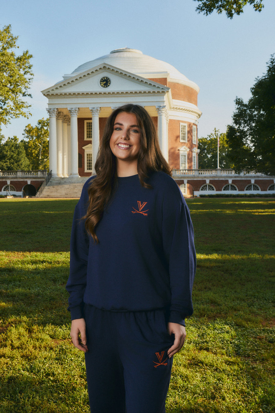 A woman stands outdoors in front of a historic domed brick building on a grassy lawn, wearing a navy blue sweatshirt and matching sweatpants with a small orange logo on the chest and thigh.