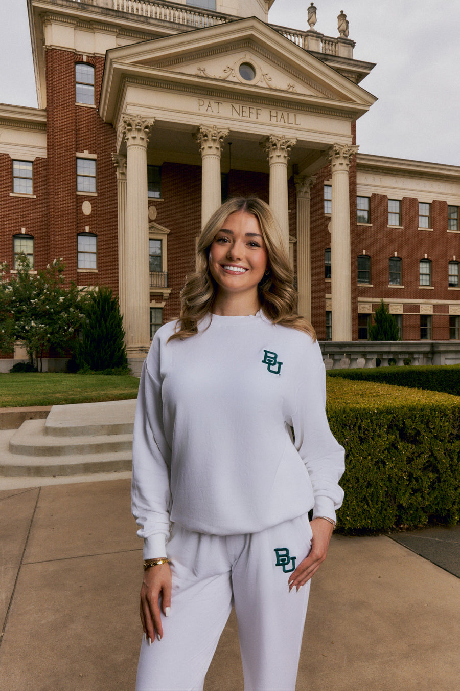 A woman stands outdoors in front of a large brick university building with columns, wearing a white sweatshirt and matching sweatpants with a green and blue 'BU' logo on the chest and thigh.