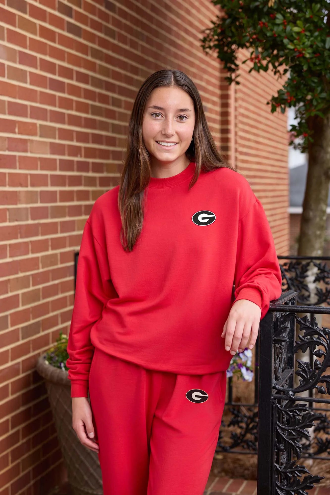 Woman wearing a red Georgia Bulldogs lounge set with matching crewneck sweatshirt and sweatpants featuring black and white “G” logo embroidery, posing outdoors against a brick wall. The coordinated collegiate outfit has a relaxed fit and casual athletic-inspired style.