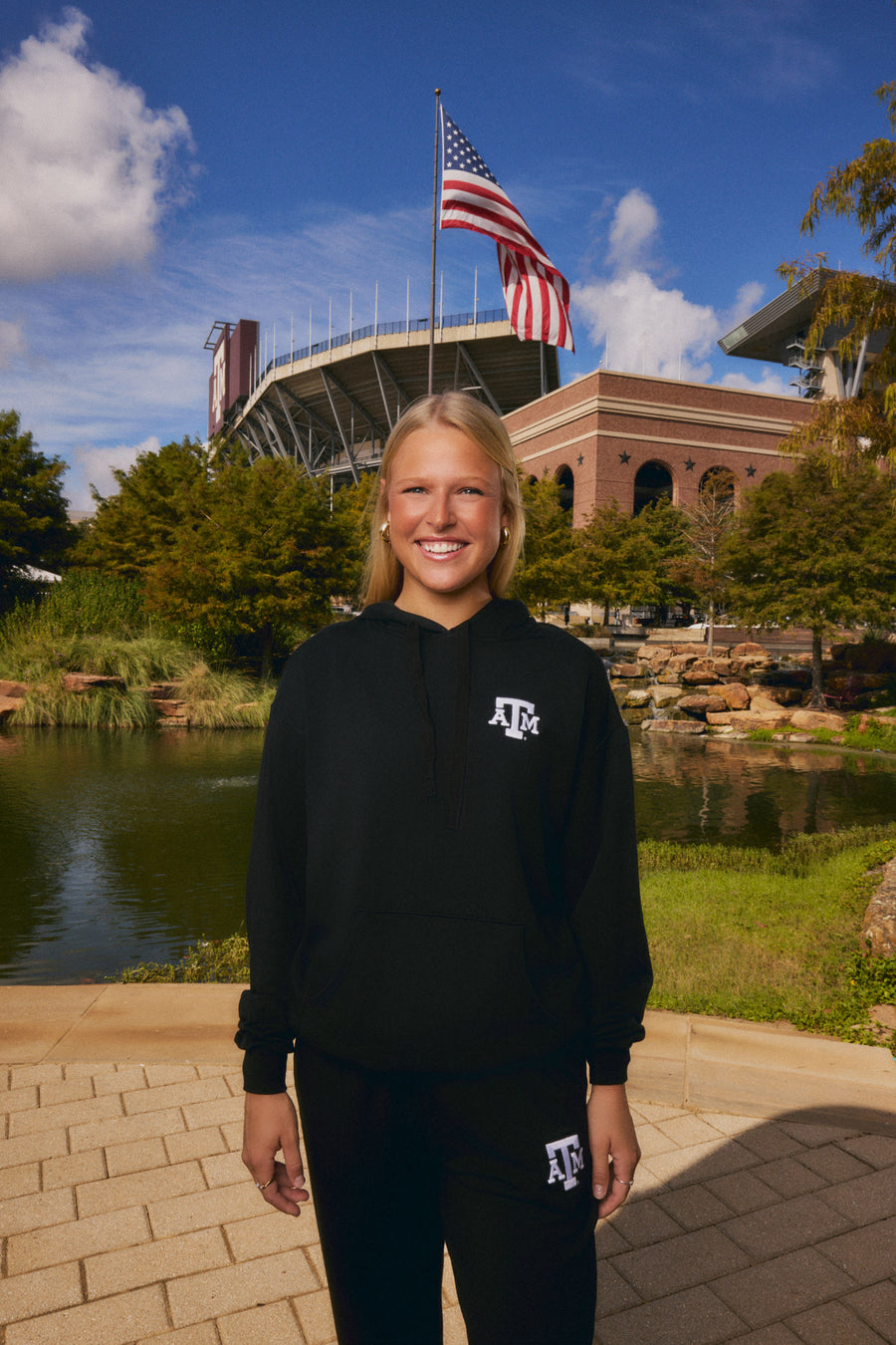 A smiling blonde woman stands outdoors in front of a stadium and pond, wearing a black hoodie and matching black sweatpants with a white A&M logo. An American flag flies above the stadium in the background.