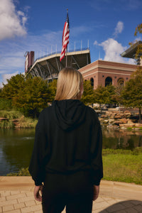 A person with blonde hair seen from behind, wearing a black hoodie and black pants, standing outdoors in front of a stadium and an American flag near a pond and brick building.