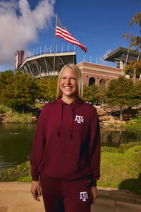 A smiling blonde woman stands outdoors in front of a stadium and pond, wearing a matching maroon hoodie and sweatpants set with a white Texas A&M logo. An American flag flies above her, with trees, grass, and blue sky in the background.