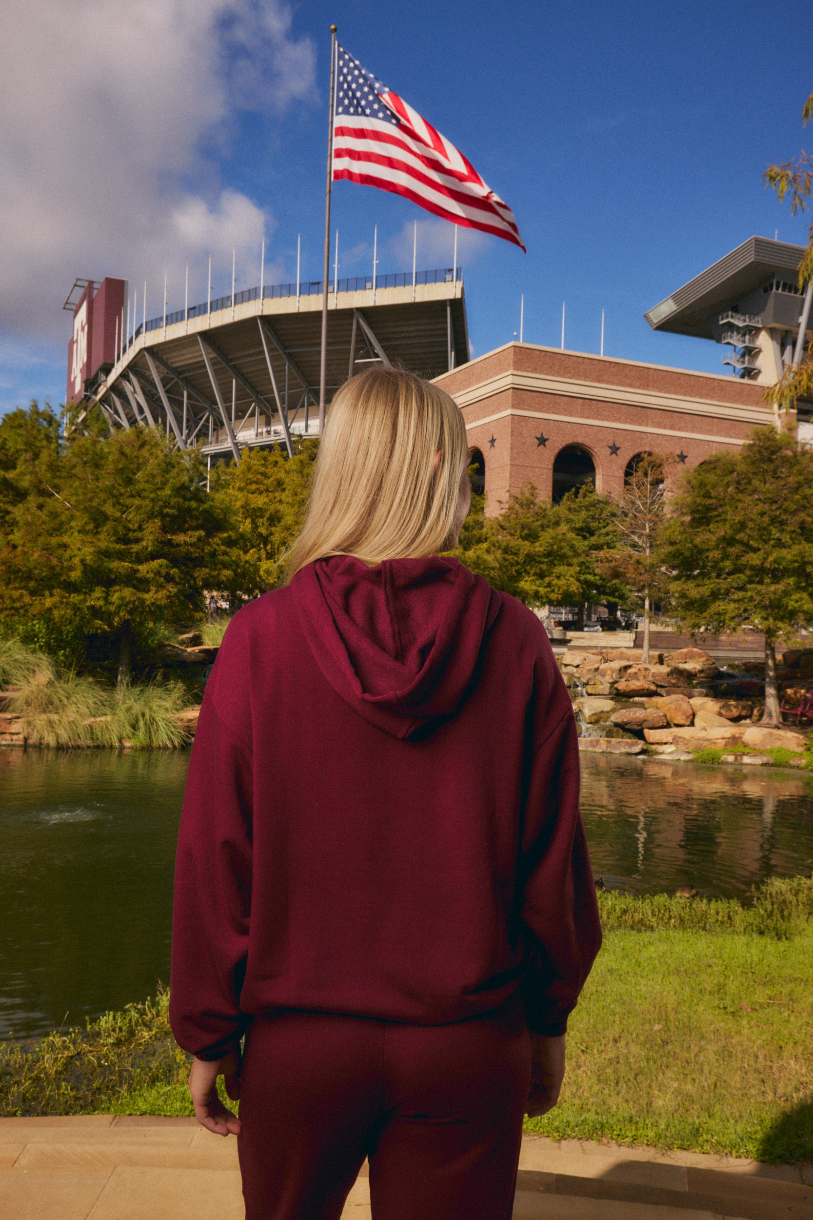 A woman viewed from behind wearing a matching burgundy hoodie and sweatpants set, standing outdoors in front of a stadium and lake with an American flag waving overhead.