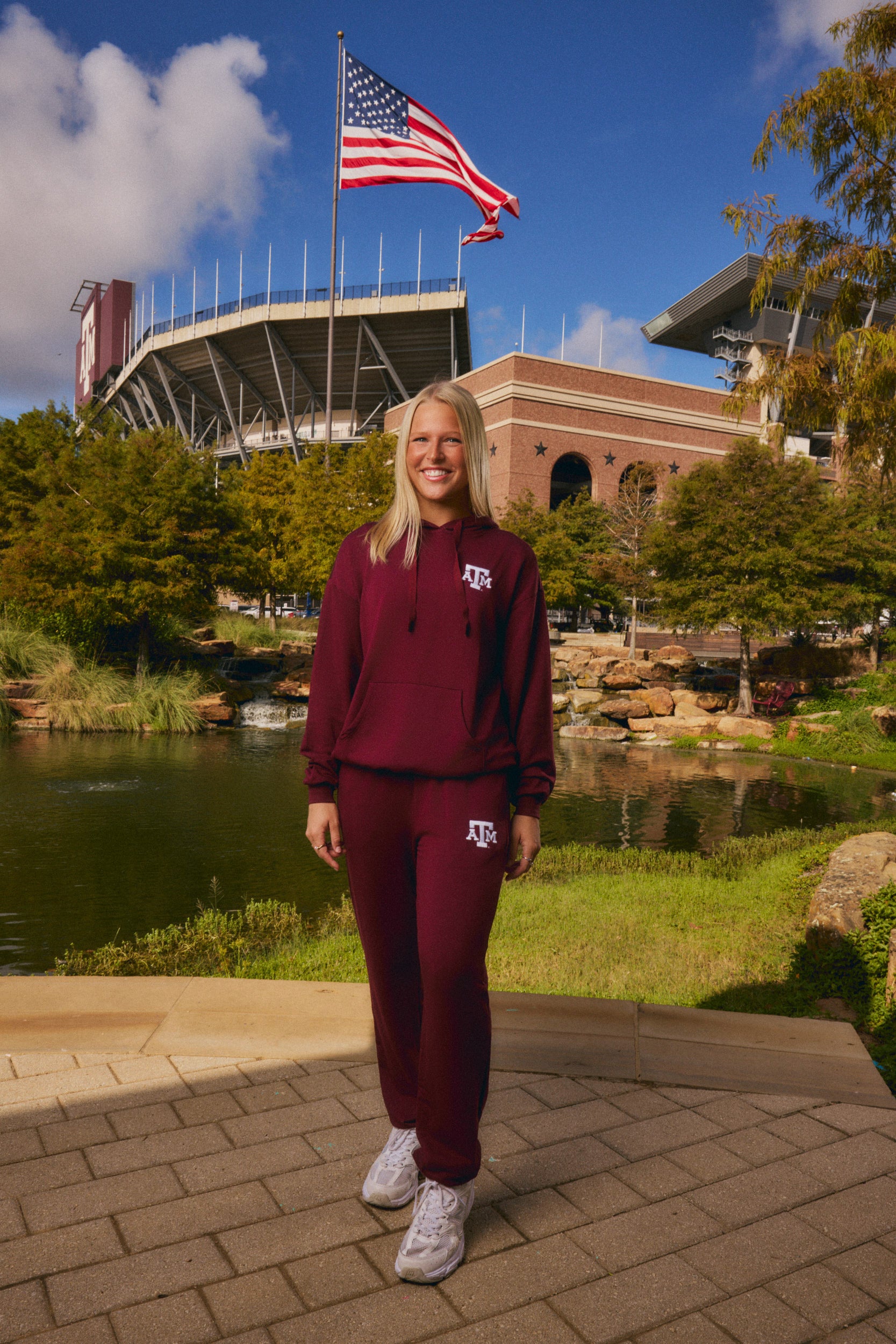 A woman stands outdoors in front of a stadium and pond, wearing a matching burgundy athletic hoodie and jogger set with white sneakers. The hoodie has a small white 'Texas' logo on the chest, and the joggers have a white 'A' logo on the thigh.