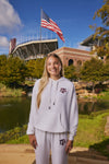 A smiling young woman stands outdoors in front of a stadium and lake, wearing a white hooded sweatshirt and matching sweatpants with a maroon Texas A&M logo on the chest and thigh.