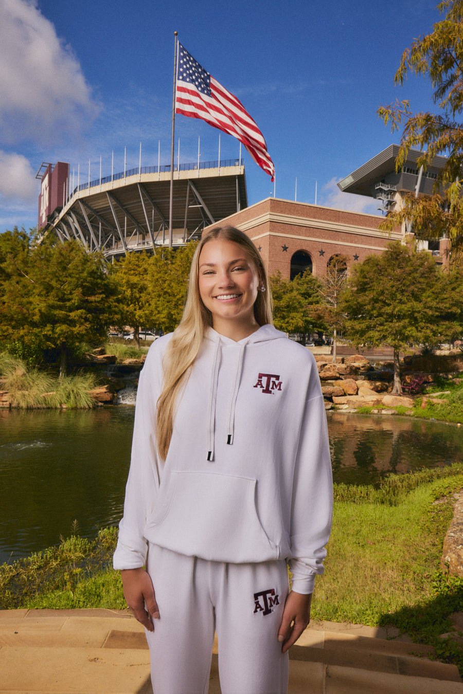 A smiling young woman stands outdoors in front of a stadium and lake, wearing a white hooded sweatshirt and matching sweatpants with a maroon Texas A&M logo on the chest and thigh.