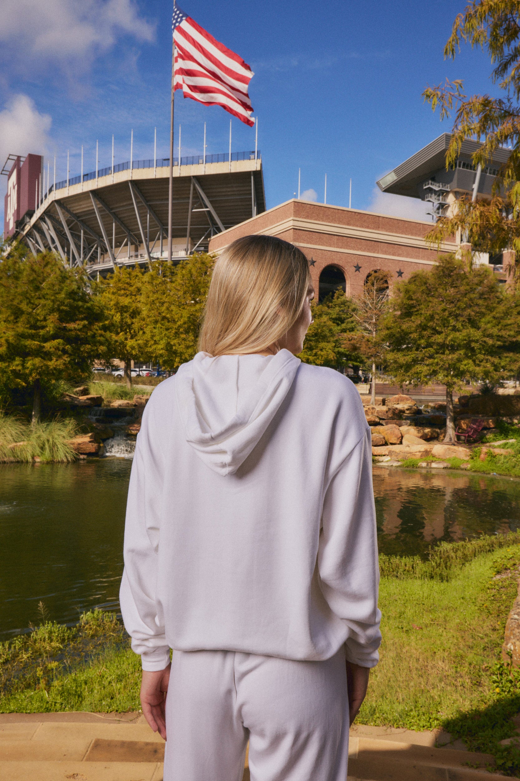 A woman with blonde hair is seen from behind, wearing a light gray hoodie and matching sweatpants, standing outdoors in front of a stadium with an American flag, trees, and a pond.