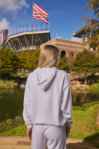 A woman with blonde hair is seen from behind, wearing a light gray hoodie and matching sweatpants, standing outdoors in front of a stadium with an American flag, trees, and a pond.