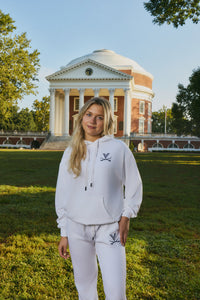 A woman stands on a grassy lawn in front of a large classical building, wearing a matching white hoodie and sweatpants set with a small navy logo on the chest and thigh.