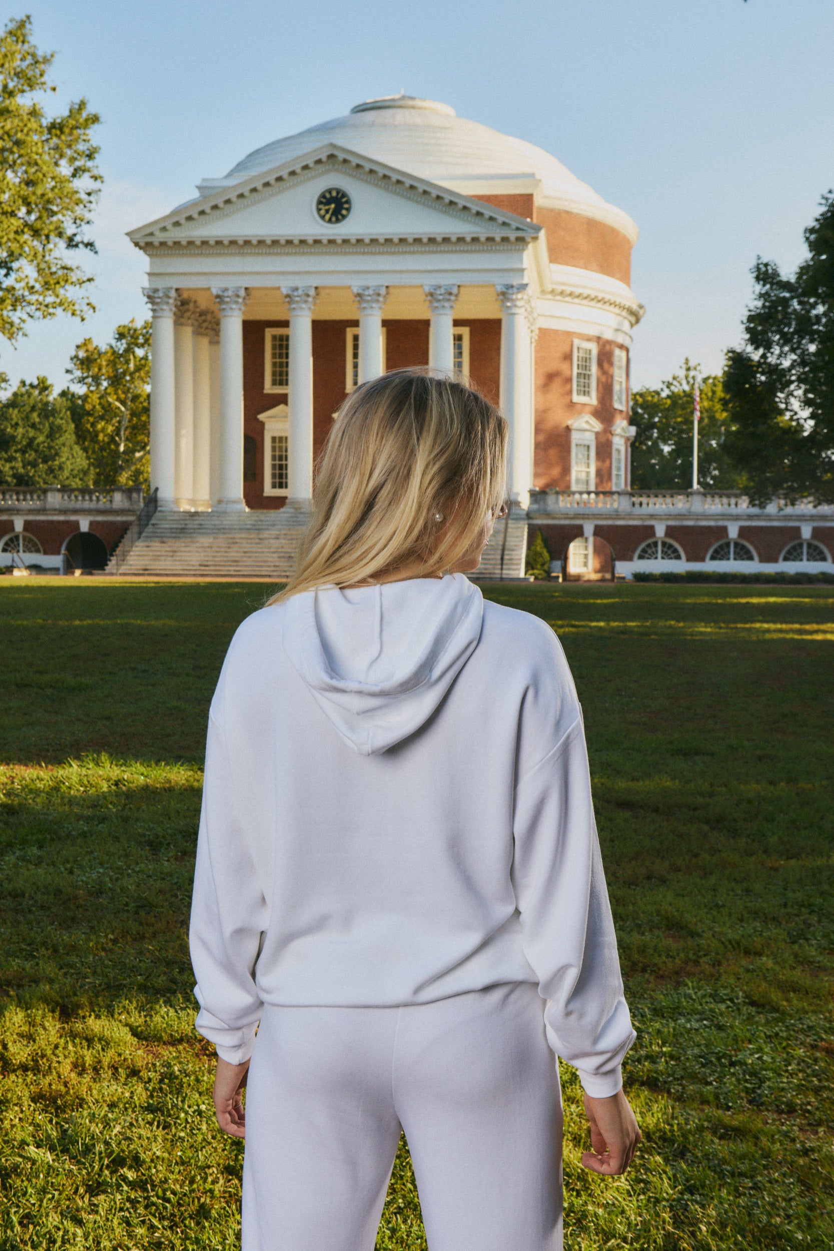 A woman viewed from behind wearing a light gray matching lounge set, standing on a grassy lawn with the historic Virginia domed capitol building in the background.
