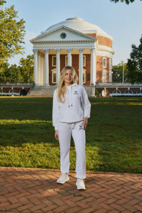 A woman stands outdoors in front of a large classical brick and white-domed building on a grassy lawn, wearing a matching white hoodie and sweatpants set with small dark embroidered logos. The photo is taken in warm natural light on a brick path.