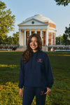 A woman stands outdoors on a grassy lawn in front of a large classical building, wearing a navy blue hoodie and matching sweatpants with a small orange embroidered logo on the chest and thigh.