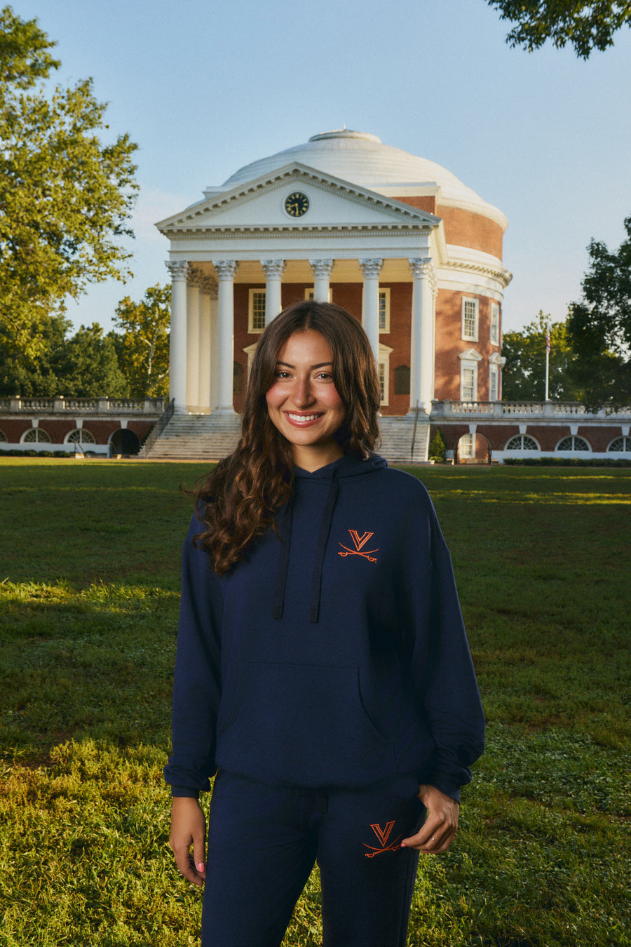 A woman stands outdoors on a grassy lawn in front of a large classical building, wearing a navy blue hoodie and matching sweatpants with a small orange embroidered logo on the chest and thigh.