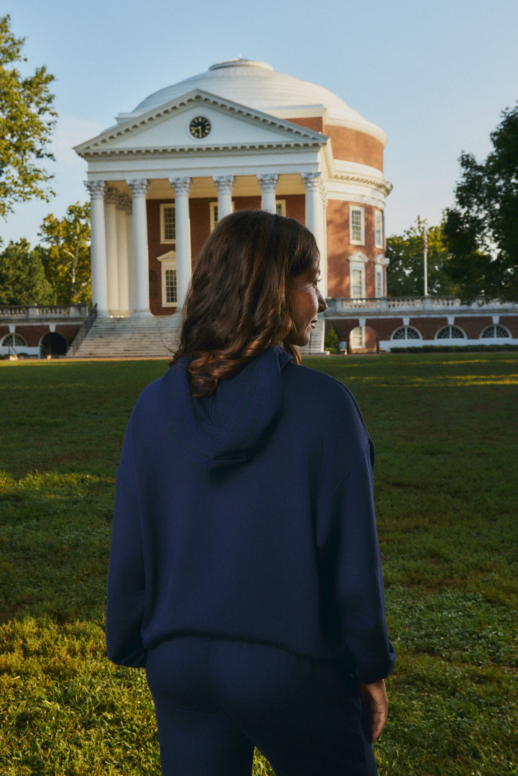 A woman wearing a navy blue hoodie and matching sweatpants stands on a grassy lawn, viewed from behind, with the historic domed building of Mount Vernon in Virginia in the background.