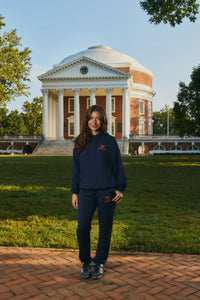 A woman stands in front of a historic red brick domed building on a sunny day, wearing a navy blue sweatshirt and matching sweatpants with a small red logo on the chest.