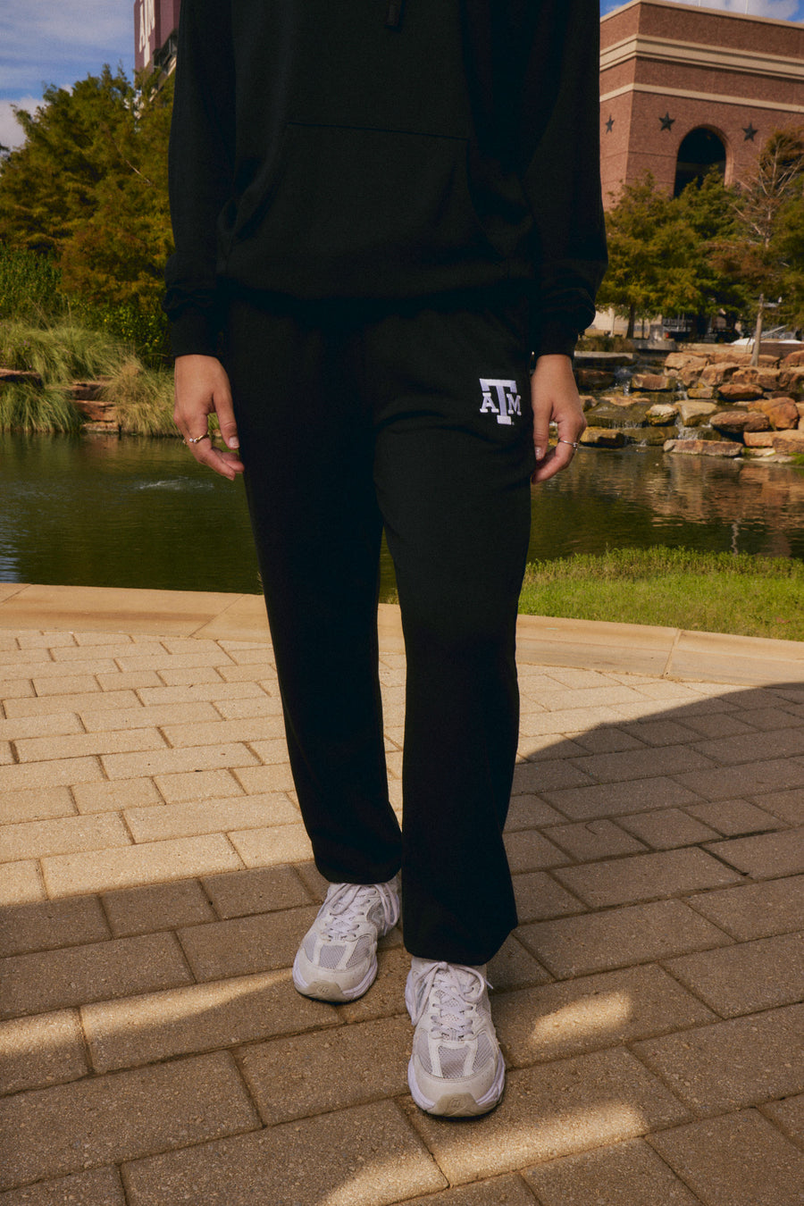 A person wearing a black hoodie and black sweatpants with a white Texas A&M logo on the left thigh, standing outdoors on a paved walkway near a pond.