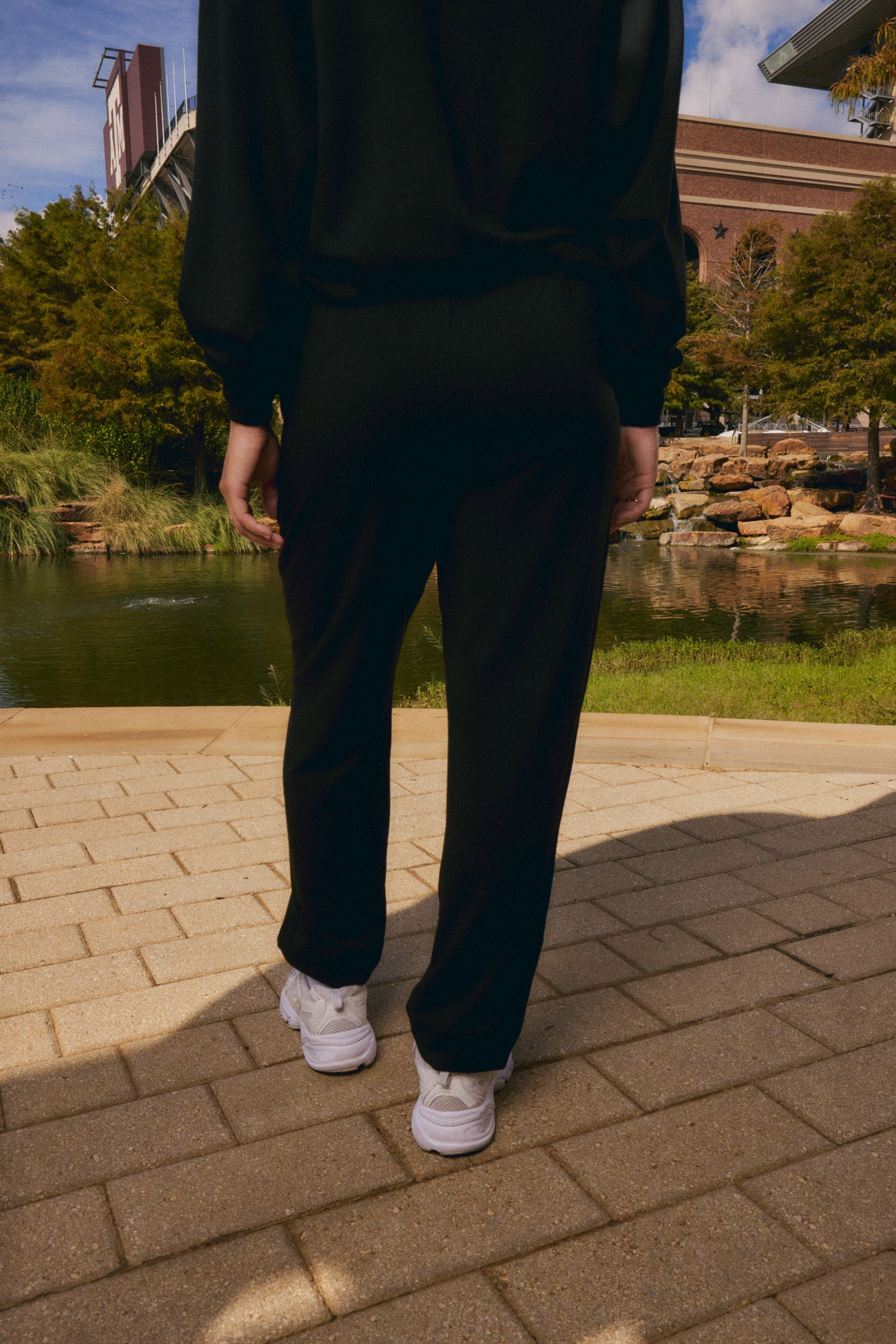 A person viewed from behind wearing a black long-sleeve top and black pants, standing on a paved walkway outdoors near a pond and trees.