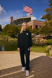 A woman stands outdoors on a brick walkway in front of a stadium and pond, wearing a matching black hoodie and sweatpants set with white logo details. The background includes trees, a pond, and a large American flag flying above the stadium.