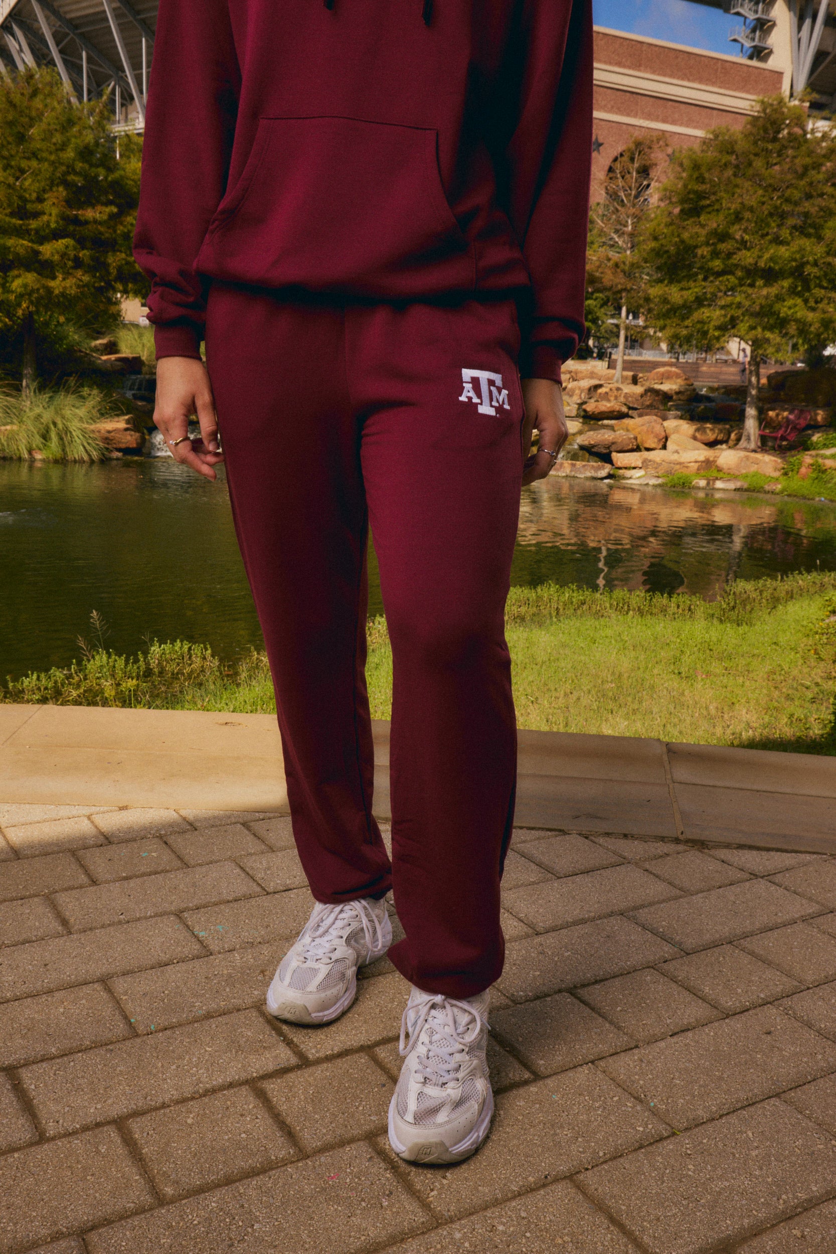 A person wearing a matching burgundy sweatshirt and jogger sweatpants set with a white 'ATK' logo on the thigh, standing outdoors on a paved walkway near a pond and stadium background.