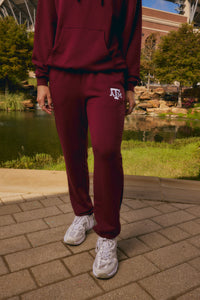 A person wearing a matching burgundy sweatshirt and jogger sweatpants set with a white 'ATK' logo on the thigh, standing outdoors on a paved walkway near a pond and stadium background.