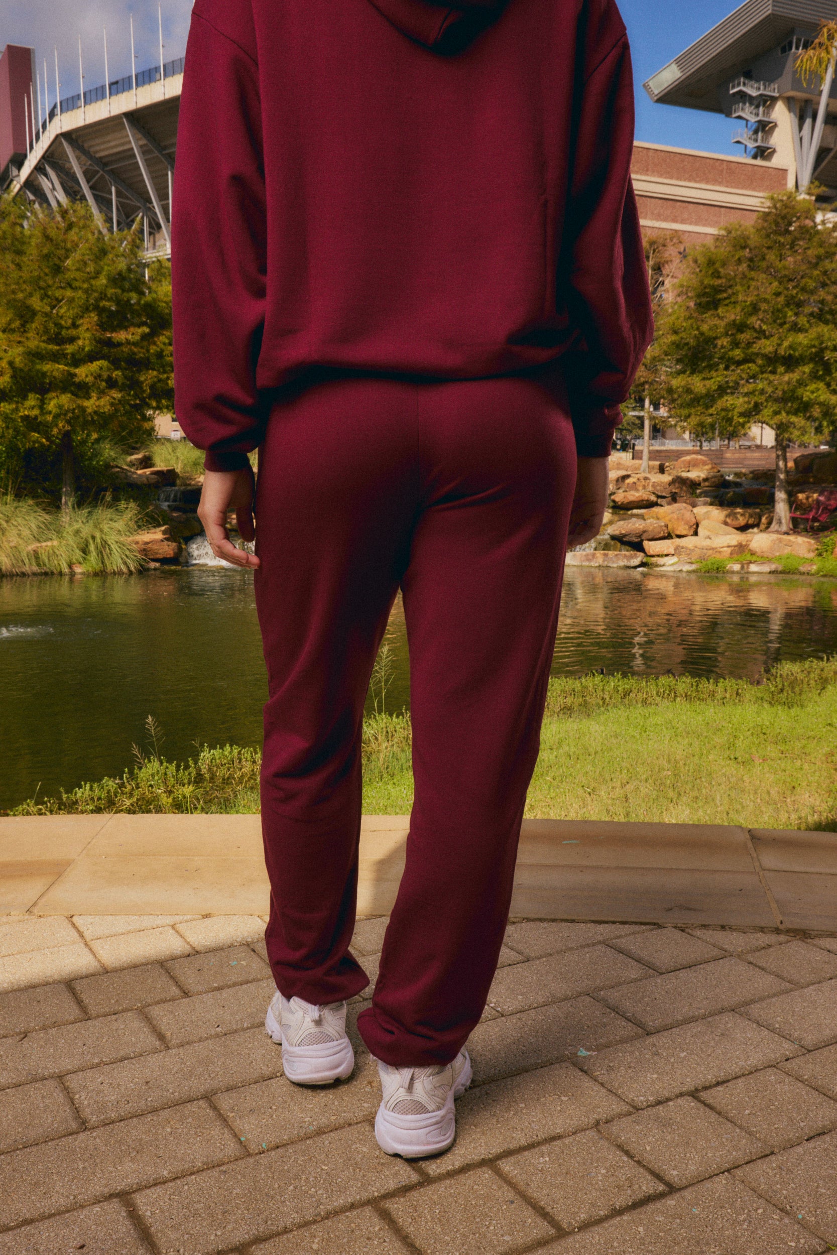 A person viewed from behind wearing a matching burgundy hoodie and jogger sweatpants set, standing on a paved walkway outdoors near a pond and trees.
