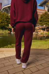 A person viewed from behind wearing a matching burgundy hoodie and jogger sweatpants set, standing on a paved walkway outdoors near a pond and trees.