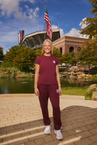 A woman stands outdoors in front of a stadium and pond, wearing a matching maroon short-sleeve top and jogger pants set with white sneakers. The set has a small white logo on the chest and thigh.