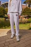 A person wearing a white matching hoodie and sweatpants set with a maroon Texas A&M logo, standing outdoors near a pond and stadium.