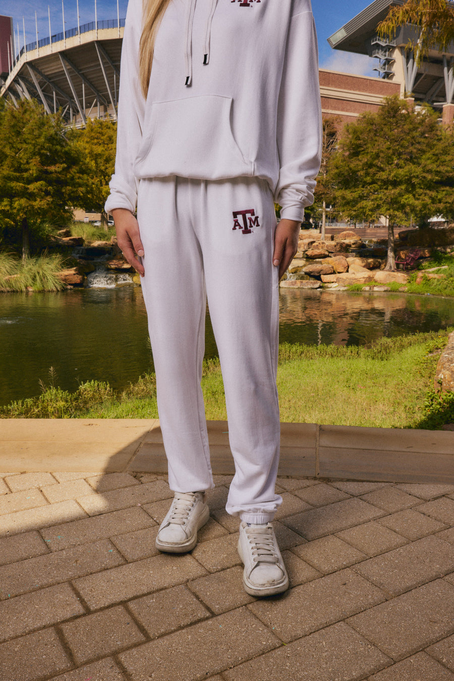 A person wearing a white matching hoodie and sweatpants set with a maroon Texas A&M logo, standing outdoors near a pond and stadium.