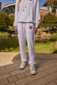 A person wearing a white matching hoodie and sweatpants set with a maroon Texas A&M logo, standing outdoors near a pond and stadium.