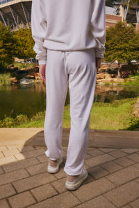 A person viewed from behind wearing a matching light off-white sweatshirt and jogger pants with white sneakers, standing on a paved walkway with a pond and greenery in the background.
