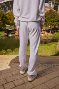 A person viewed from behind wearing a matching light off-white sweatshirt and jogger pants with white sneakers, standing on a paved walkway with a pond and greenery in the background.