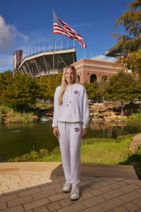 A woman stands outdoors in front of a stadium and pond, wearing a white matching sweatshirt and sweatpants set with Texas A&M athletic logos. An American flag is visible in the background under a blue sky.