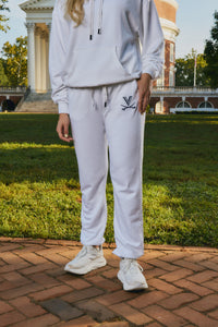 A person wearing a matching white sweatshirt and jogger pants set with white sneakers, standing outdoors on a brick path with grass and a classical building in the background.