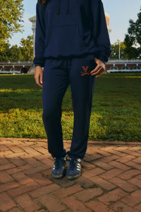 A person wearing a navy blue sweatshirt and matching sweatpants stands outdoors on a brick patio with grass and trees in the background. The sweatpants have a small orange 'V' logo on the upper left thigh, and the person is wearing dark blue sneakers.