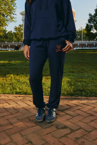A person wearing a navy blue sweatshirt and matching sweatpants stands outdoors on a brick patio with grass and trees in the background. The sweatpants have a small orange 'V' logo on the upper left thigh, and the person is wearing dark blue sneakers.