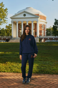 A woman stands outdoors in front of a large classical brick building on a grassy lawn, wearing a navy blue matching hoodie and sweatpants set with a small red logo on the chest and thigh.