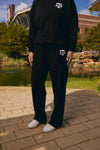 A person wearing a black sweatshirt and matching black sweatpants with white Texas A&M University 'AT&M' logo graphics on the chest and thigh, standing outdoors on a paved walkway near a pond and stadium.