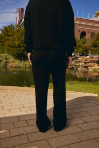 A person viewed from behind wearing a black long-sleeve top and black wide-leg pants, standing on a paved walkway outdoors near a pond and brick building.
